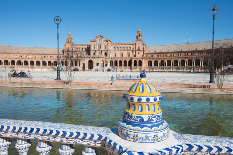 Blue-and-yellow ceramic finial and tiled balustrade beside the Plaza de España canal, with central pavilion across the water.
