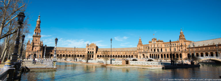Curving canal, tiled bridges, arcaded façades, and a tall brick tower at Plaza de España under a clear blue sky.