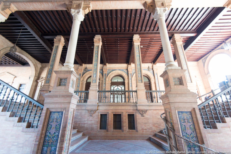 Grand interior staircase at Plaza de España with pale columns, colorful tile panels, iron railings, and a dark timber ceiling.