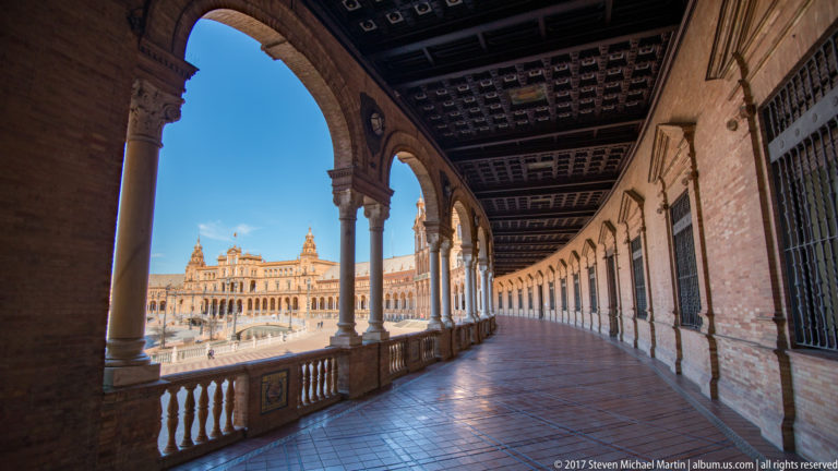 Curving upper arcade at Plaza de España with columns, tiled floor, coffered ceiling, and sunlit pavilion beyond.