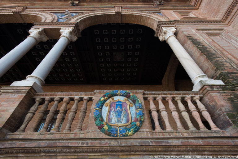 Round multicolored ceramic heraldic medallion set in a brick balustrade beneath columns at Plaza de España.