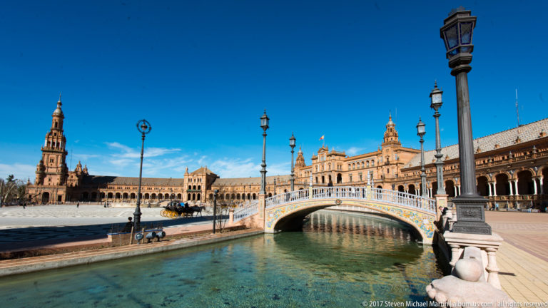 Low ceramic-tiled bridge crosses the Plaza de España canal with brick towers, arcades, and ornamental lamps beyond.