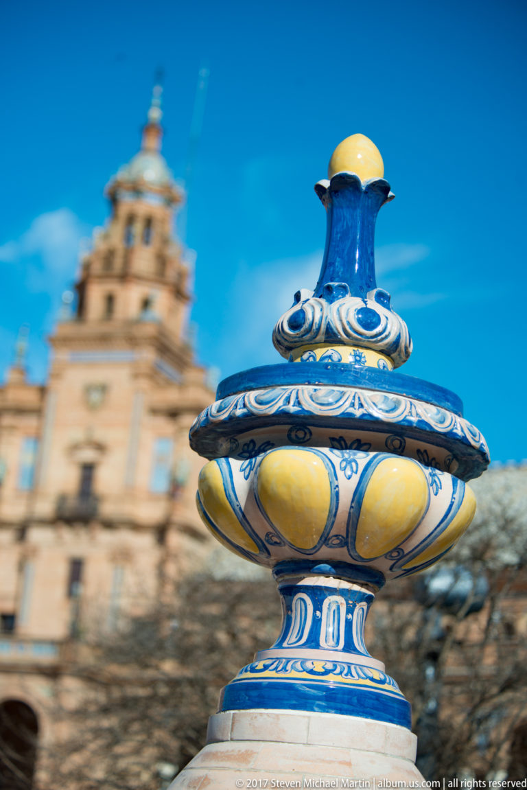 Blue-and-yellow glazed ceramic urn finial in sharp focus with a Plaza de España pavilion blurred in the background.