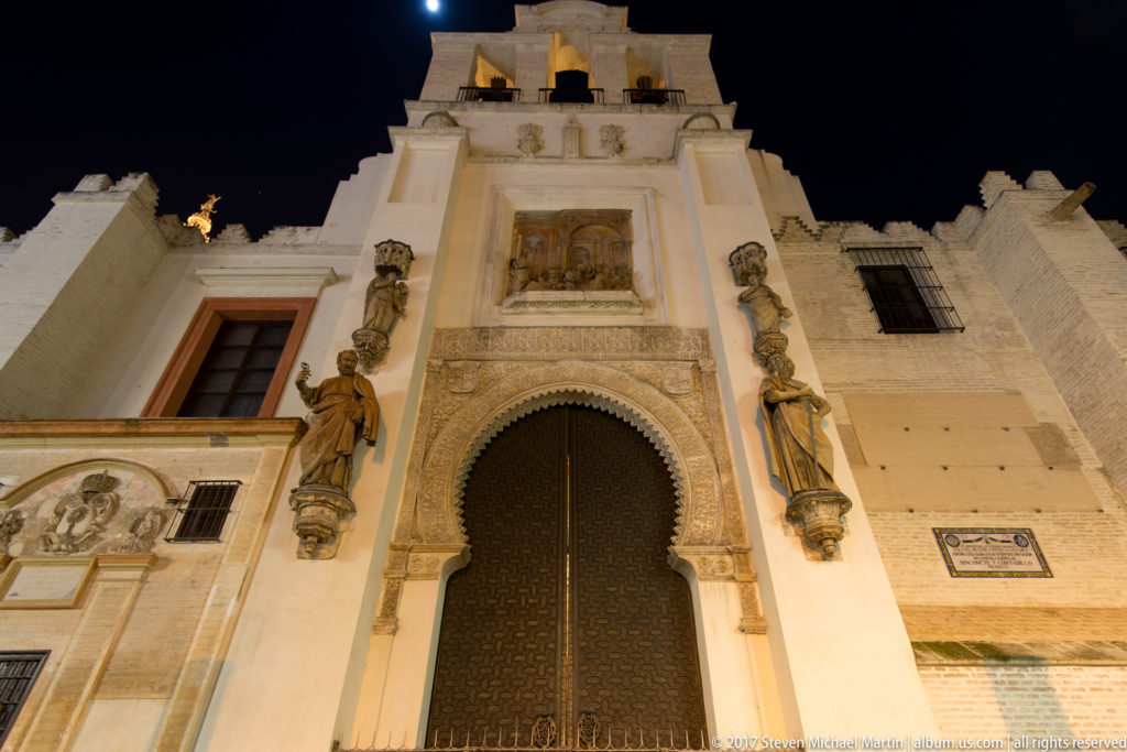 Tall arched cathedral doorway with dark doors, flanking statues, relief sculpture, and warm nighttime lighting.