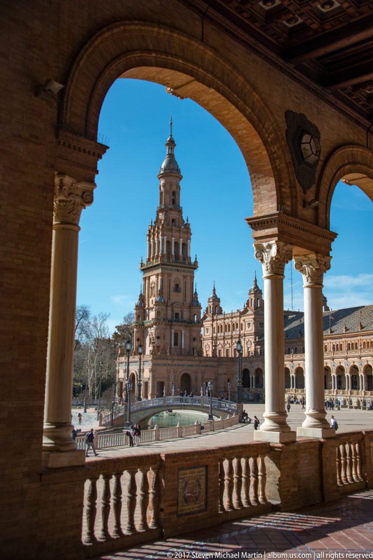 Sunlit Plaza de España pavilion and tower framed by the shaded arches, columns, and balustrade of an upper gallery.