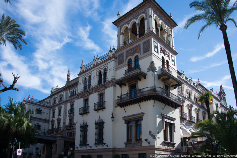 Ornate white hotel façade with arched tower, balconies, decorative panels, and palm trees against blue sky.