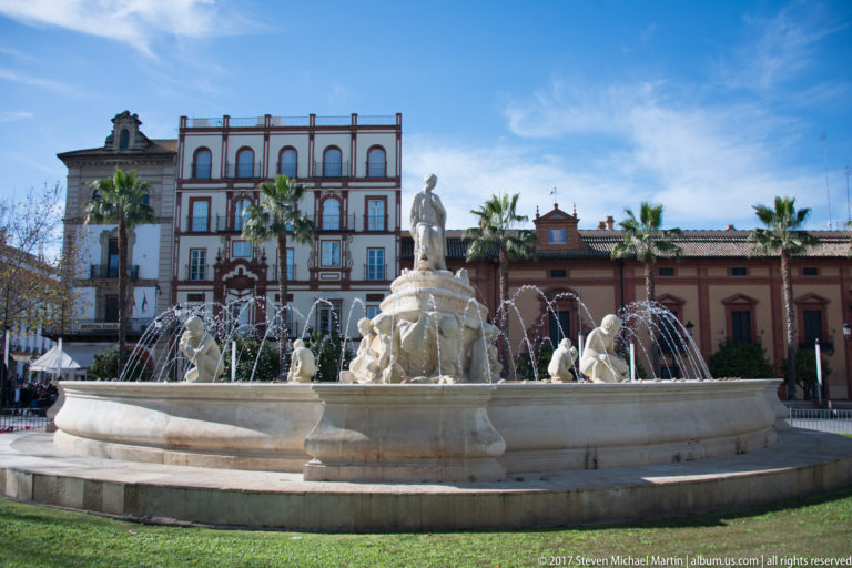Large circular fountain with standing female statue, sculpted figures, arcing jets, historic buildings, and palms.