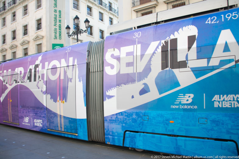 Articulated blue and purple sightseeing vehicle labeled SEVILLA parked along a city street.