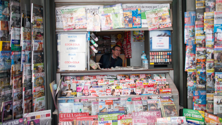 Vendor inside crowded newsstand surrounded by Spanish newspapers, magazines, drinks, snacks, and tobacco displays.