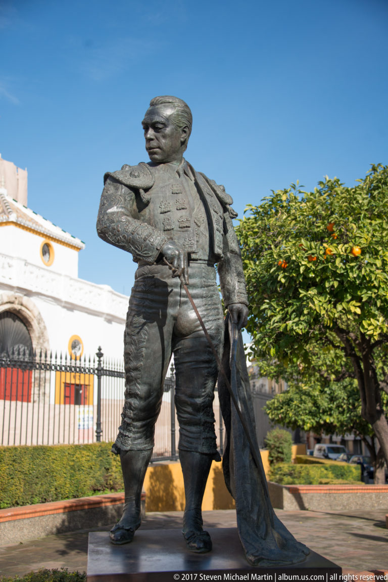 Bronze statue of a matador holding a cape beside white-and-ochre bullring buildings and iron fencing.