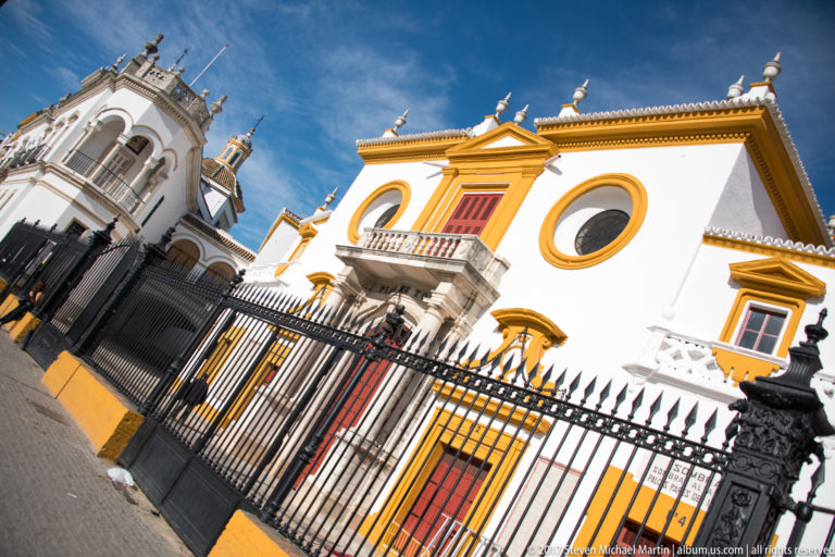 Tilted street view of white-and-ochre Real Maestranza façade behind black iron fencing and red doors.