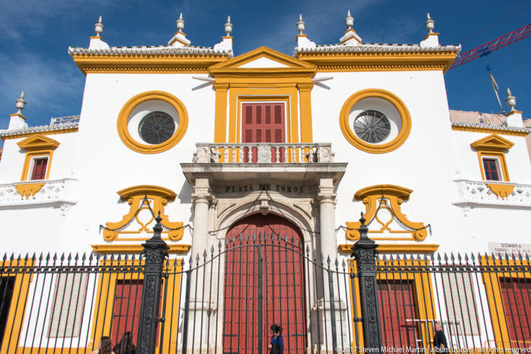 Frontal white-and-ochre bullring façade with red central gate, round windows, stone portal, and black iron fence.