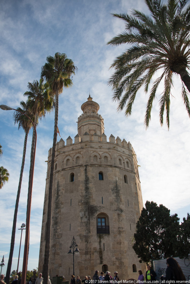 Pale stone Torre del Oro framed by tall palms, with crenellations and its smaller upper tower visible.
