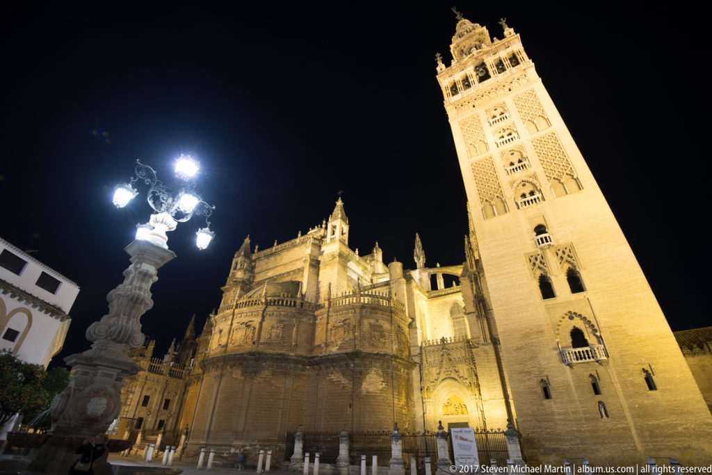 Seville Cathedral and illuminated Giralda at night, with the Gothic apse and an ornate plaza lamp in view.