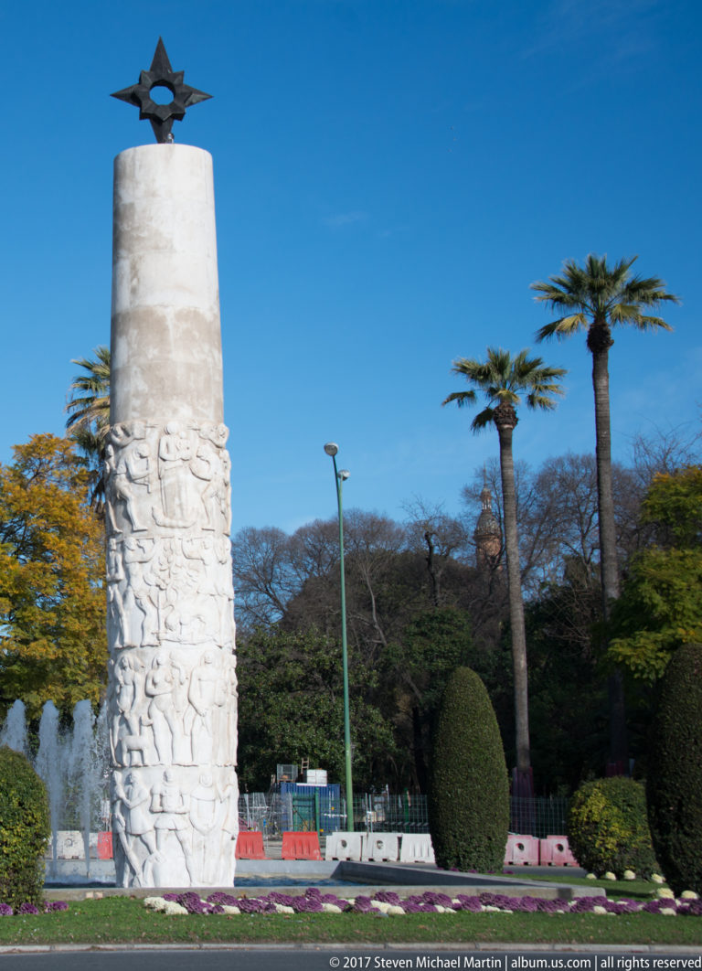 White monument column with carved relief figures standing among palms, clipped shrubs, flowers, and a park fountain.