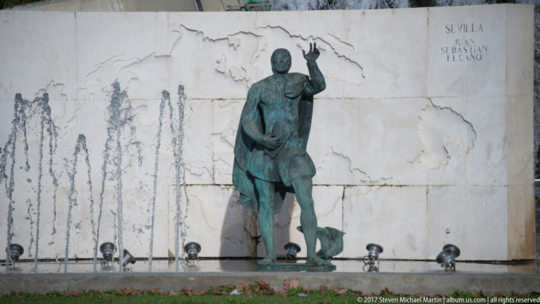 Bronze Juan Sebastián Elcano figure before inscribed stone wall and a row of fountain jets.