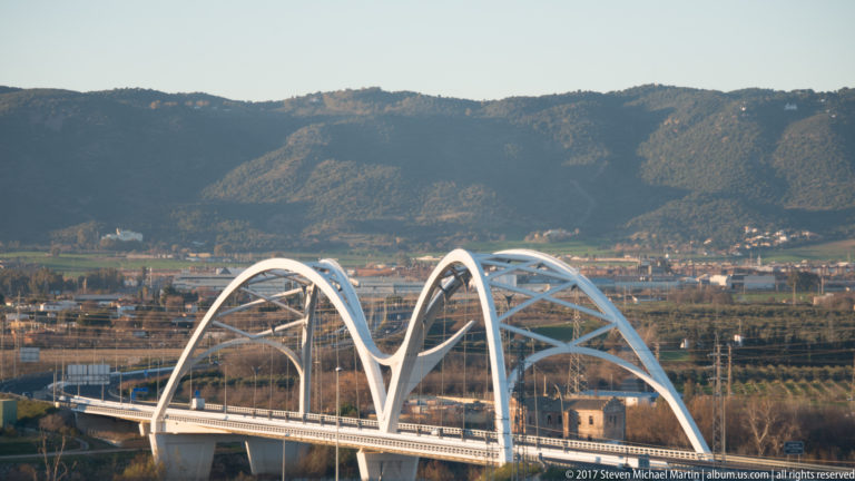 Modern white road bridge with sweeping steel arches crossing lowlands beneath wooded hills outside Seville.