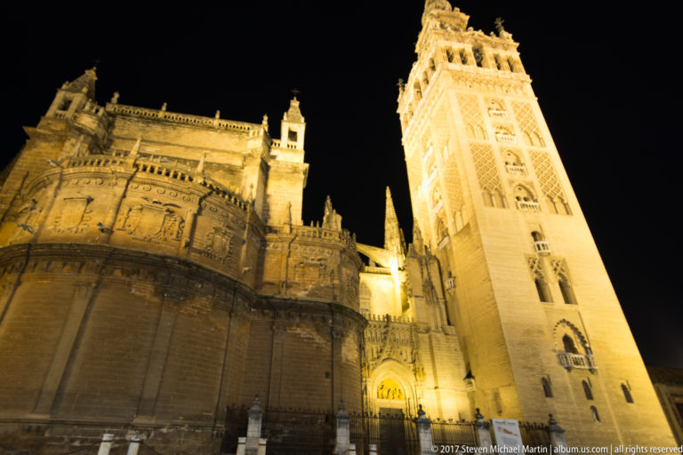 Low-angle night view of the illuminated Giralda beside the massive Gothic apse of Seville Cathedral.