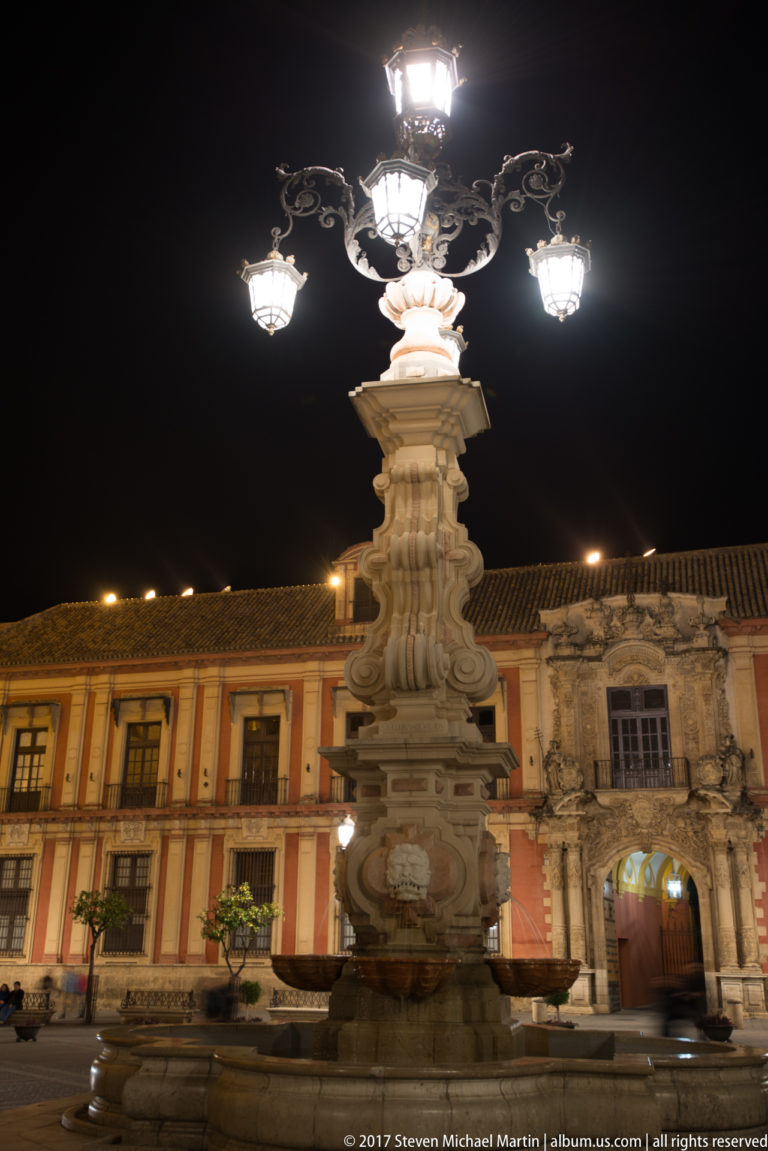 Ornate multi-lamp fountain in Plaza Virgen de los Reyes before the red façade of the Archbishop’s Palace.