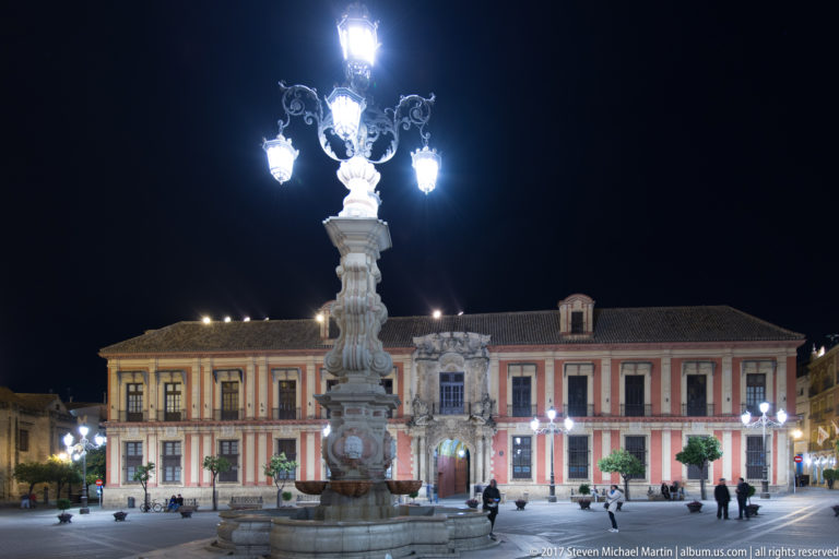 Wide night view of Plaza Virgen de los Reyes with its ornate fountain-lamp, Archbishop’s Palace, trees, and pedestrians.