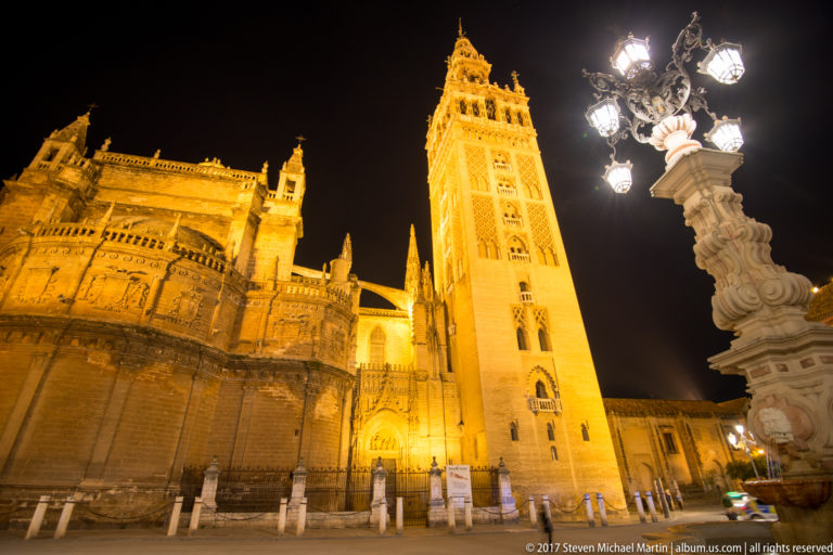 Seville Cathedral and Giralda illuminated at night with the Plaza Virgen de los Reyes fountain-lamp at right.