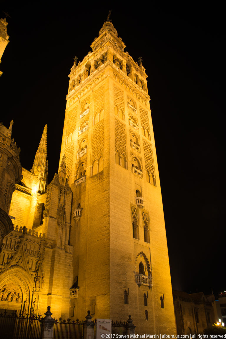 Close low-angle night view of the illuminated Giralda, with part of Seville Cathedral visible at left.