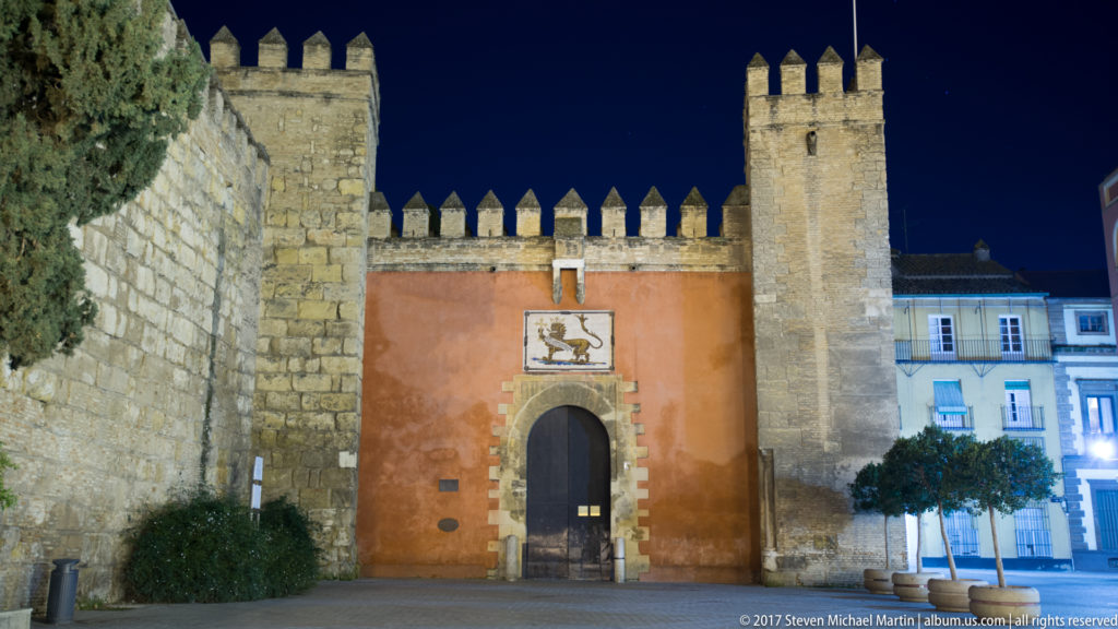 Puerta del León at the Real Alcázar, with red wall, arched doorway, lion tile, crenellations, and towers at night.