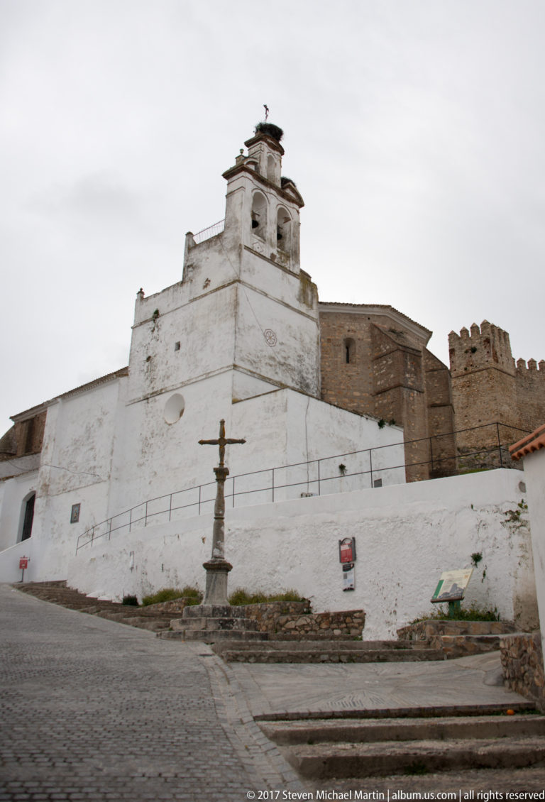White church with bell gable, stone cross, and crenellated fortress walls rising behind it.