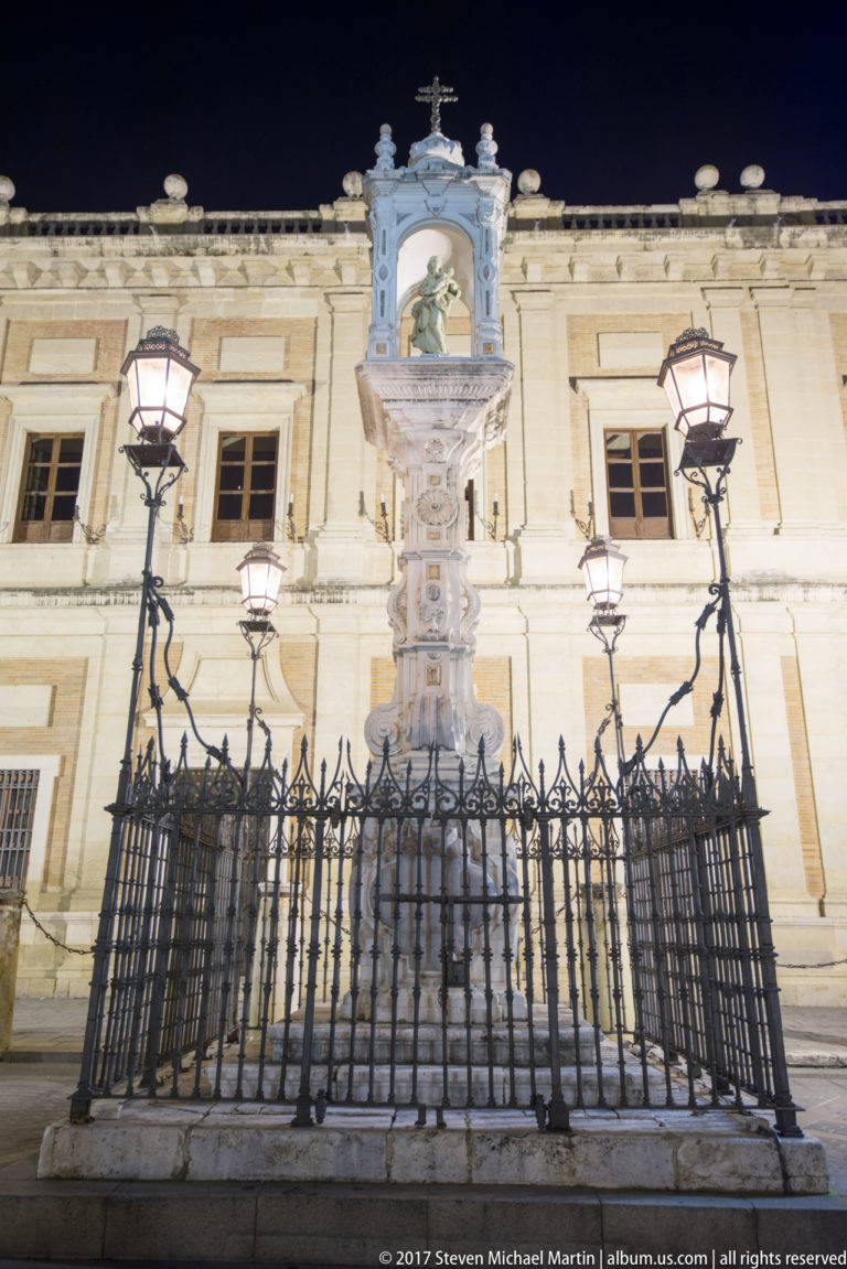 Tall carved stone cross enclosed by an iron fence and flanked by street lamps in a Seville square at night.