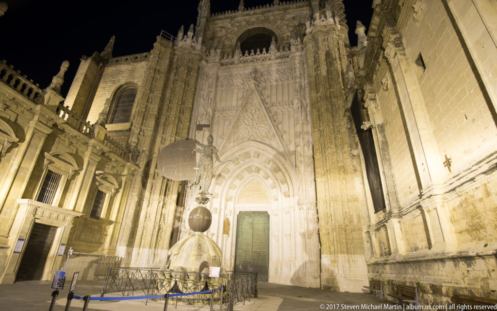 Gothic portal of Seville Cathedral illuminated at night behind a freestanding monument with a dark spherical element.