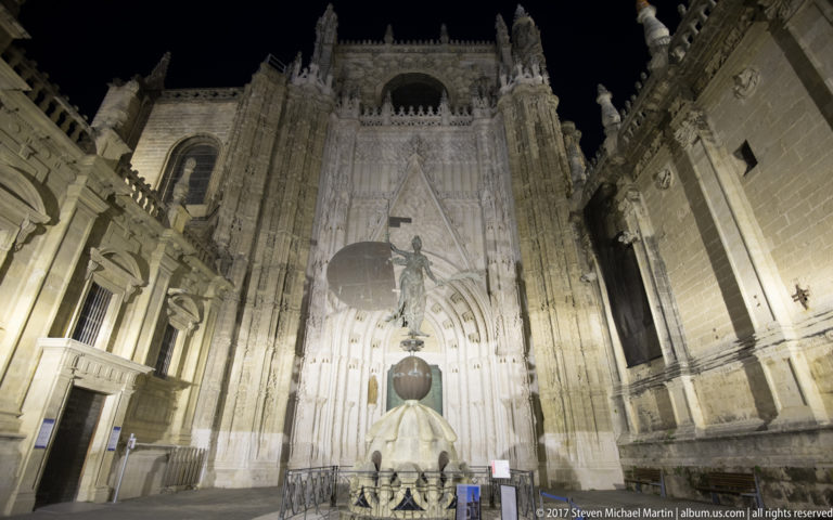 Vertical night view of a Seville Cathedral portal with a pale monument and dark sphere aligned in the foreground.