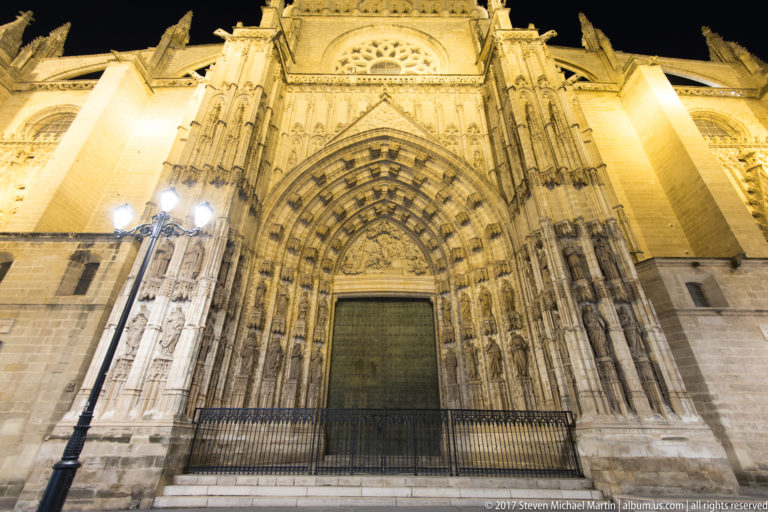 Large Gothic cathedral portal with carved archivolts, statues, tympanum, and dark central doors illuminated at night.