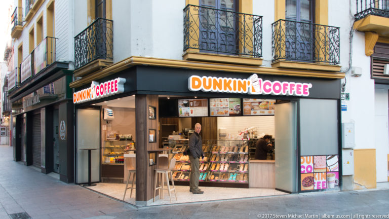 Dunkin’ Coffee corner storefront beneath wrought-iron balconies, with customers and counters visible inside.