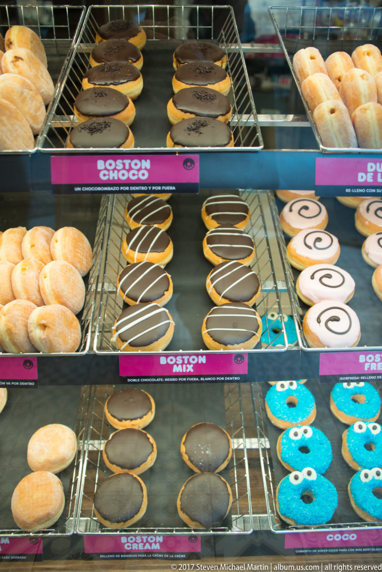 Rows of assorted doughnuts on metal racks, including chocolate, striped, spiral, powdered, and bright blue iced varieties.