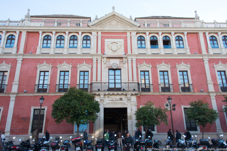 Red-and-cream historic building with central Cajasol entrance, arched windows, orange trees, scooters, and pedestrians.