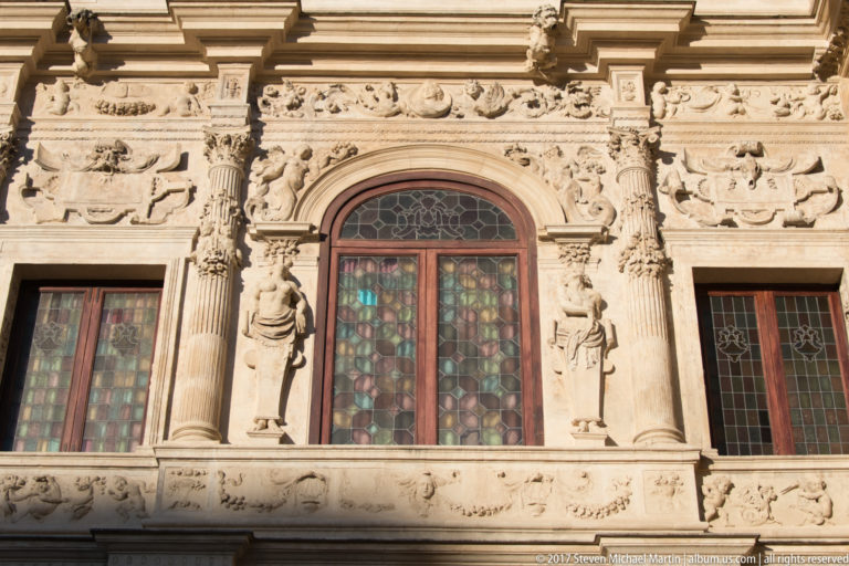 Ornate carved stone window with slender columns, relief figures, decorative frieze, and colored leaded glass.