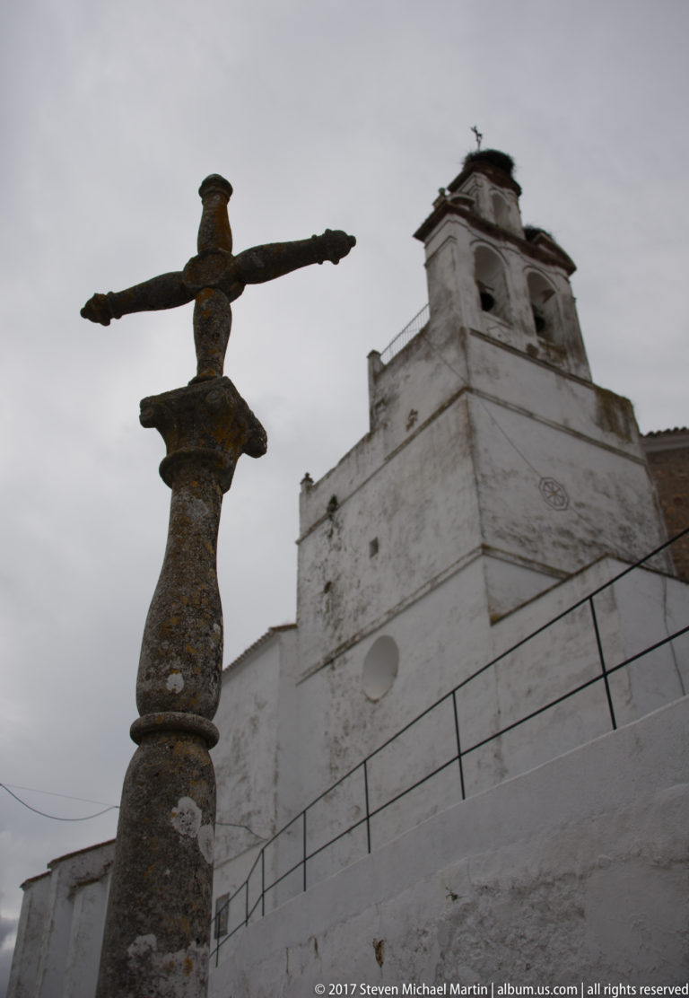 Weathered stone cross in the foreground with a white church bell gable rising behind it.