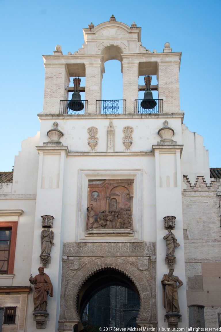 Ornamented Seville entrance with arched doorway, religious relief, sculpted figures, heraldic motifs, and two bells above.