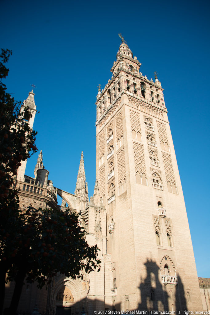 Low-angle view of the Giralda’s pale stone tower rising above Seville Cathedral beneath a deep blue sky.