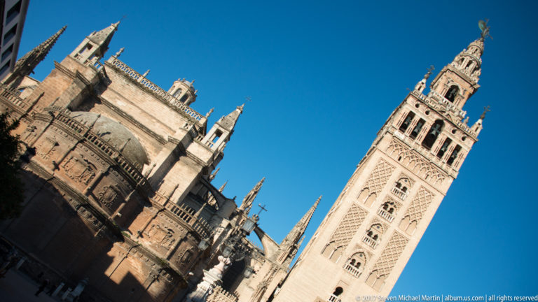 Tilted exterior view of Seville Cathedral’s Gothic masonry beside the tall Giralda tower under a clear blue sky.