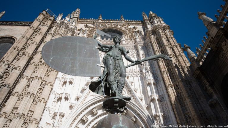 Bronze standing figure with staff seen from below against an elaborate carved Gothic portal of Seville Cathedral.
