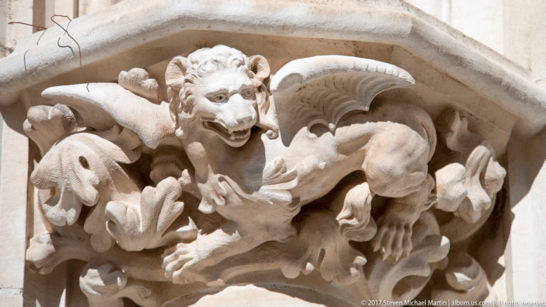 Close-up of a carved winged beast with open mouth, claws, curled tail, and foliage on pale cathedral stone.