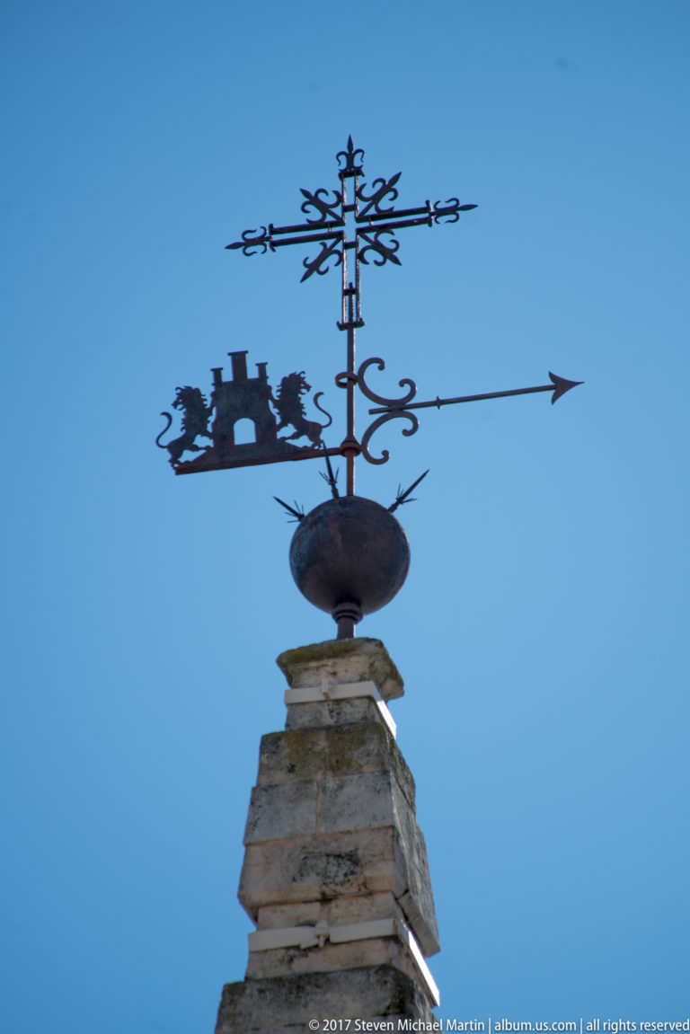 Decorative iron weather vane with arrow, compass letters, scrollwork, and heraldic silhouette atop a stone pinnacle.