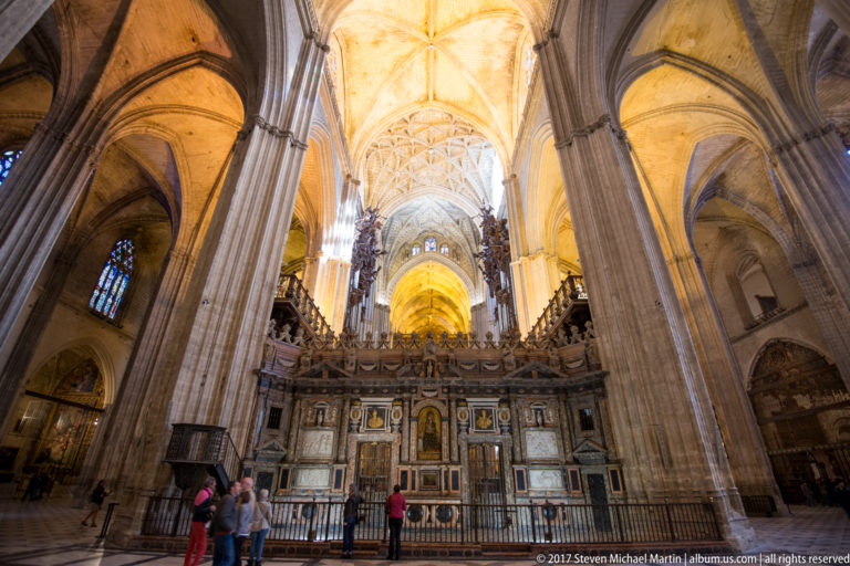 Interior of Seville Cathedral with towering piers, ribbed vaults, ornate choir enclosure, and visitors below.
