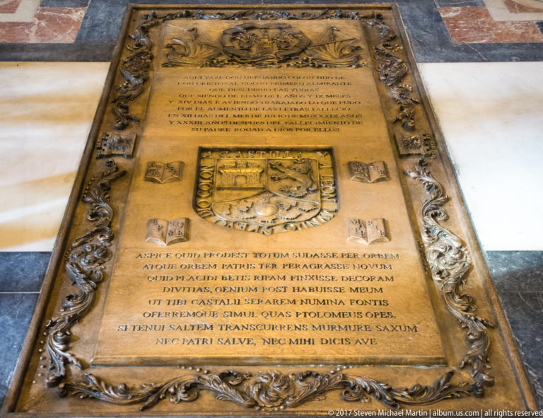 Rectangular bronze memorial slab with Latin inscription, heraldic emblems, and ornate relief border set into a stone floor.