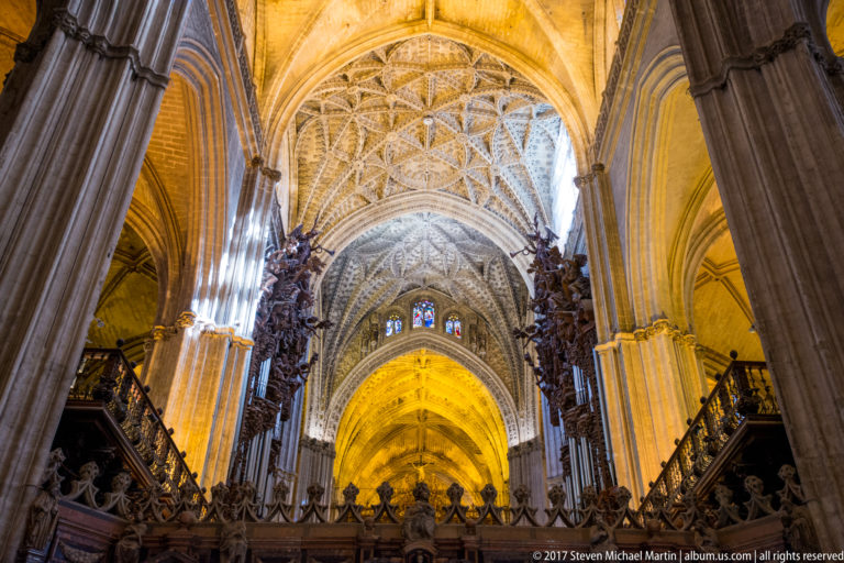 Upward view through Seville Cathedral showing clustered piers, intricate ribbed vaults, and dark carved choir screens.