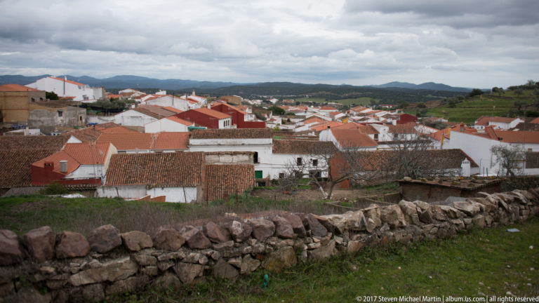 Panoramic view over white houses and red-tiled roofs toward low green hills beneath cloudy skies.