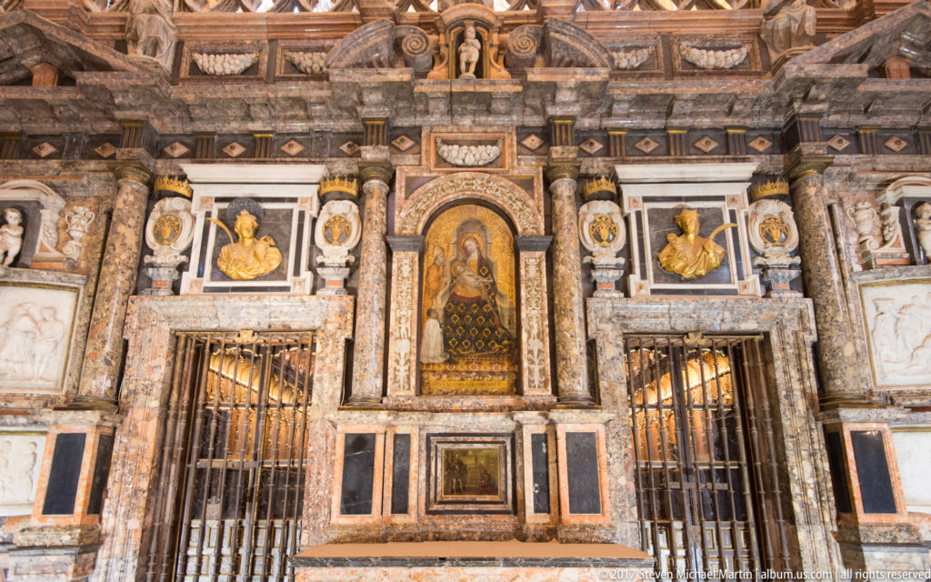 Ornate cathedral chapel wall with central devotional image, marble panels, gilded details, busts, and metal grilles.