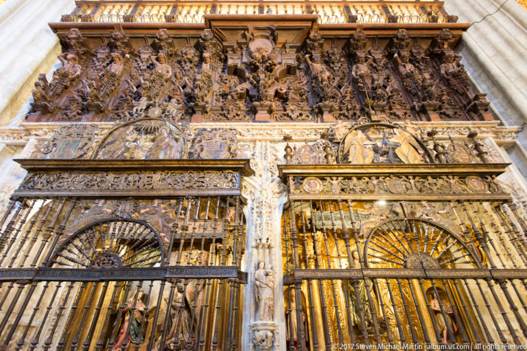 Elaborate paired cathedral grille with radiating iron bars, gilded details, carved reliefs, and sculpted figures above.