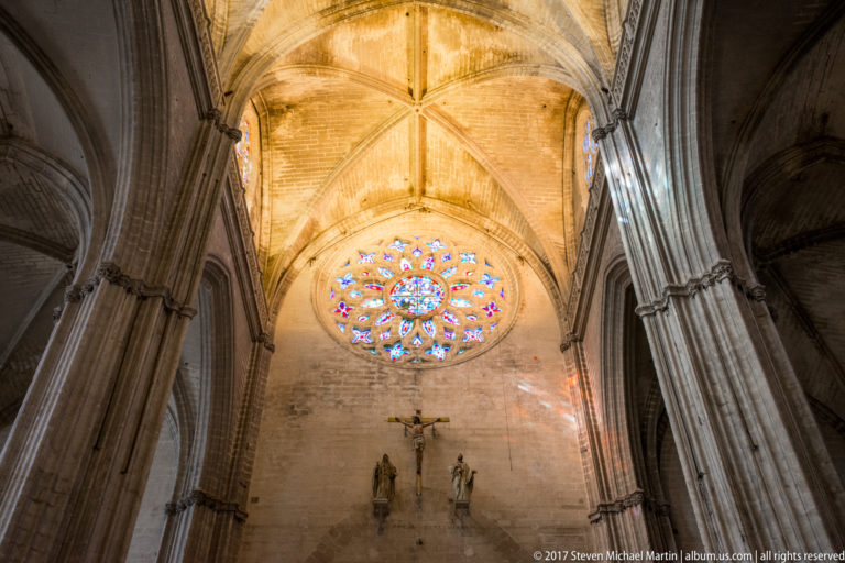 Circular stained-glass rose window above a crucifix and small figures between massive stone piers in Seville Cathedral.